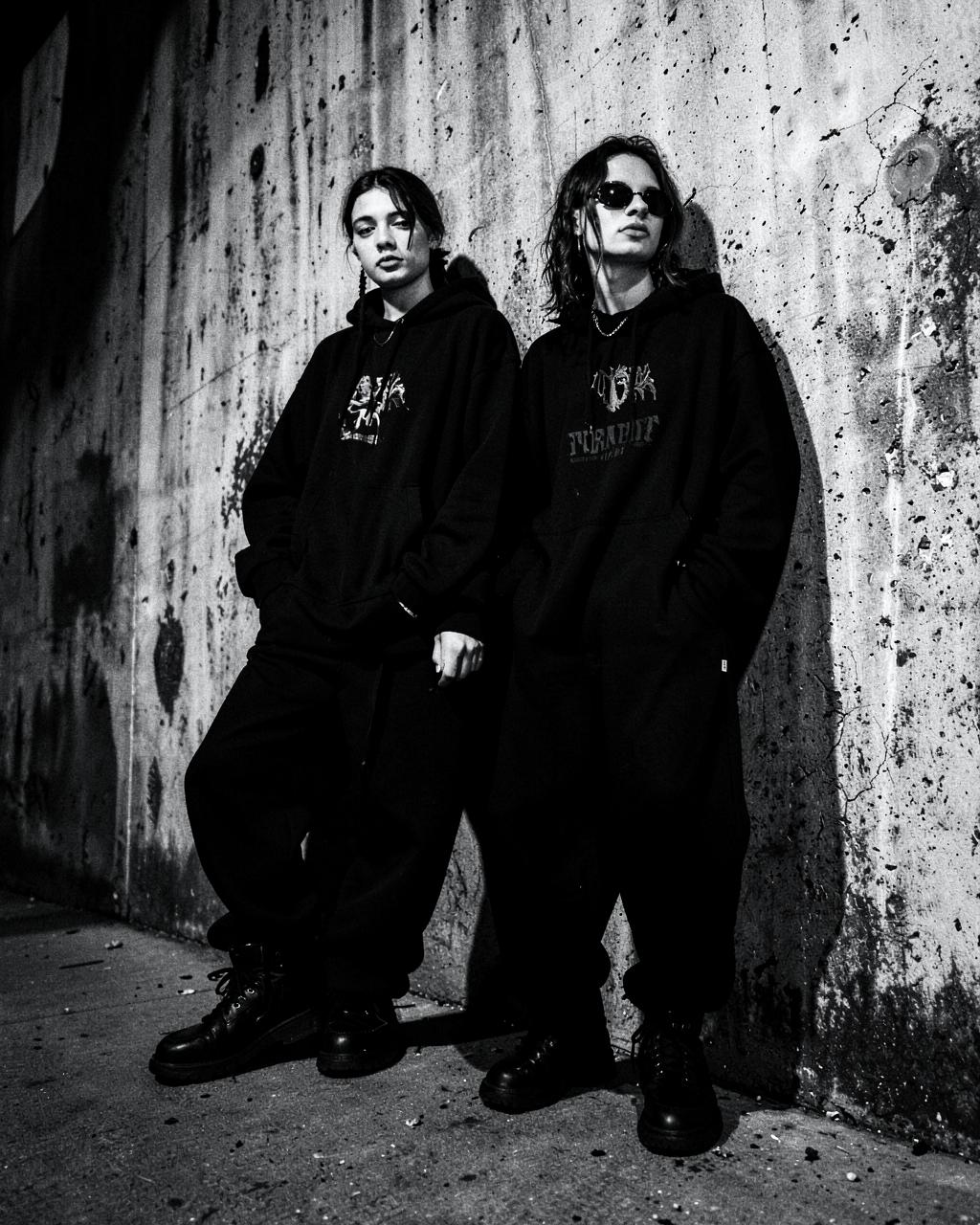 Two people in black gothic streetwear against a concrete wall